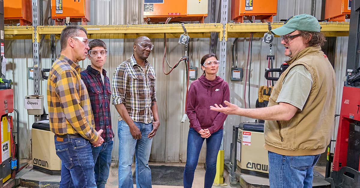 Group of diverse workers receiving safety instructions in an industrial warehouse setting, all wearing safety glasses