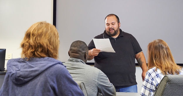 Person at front of business classroom looking at paper instructing a group of people seated at a tables