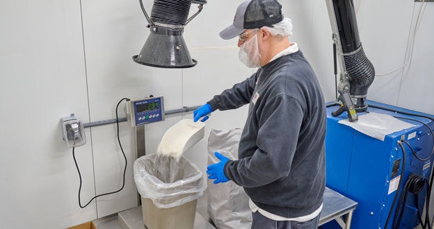 Worker scooping dry ingredients into a bagged bucket sitting on a scale with overhead exhaust sytem
