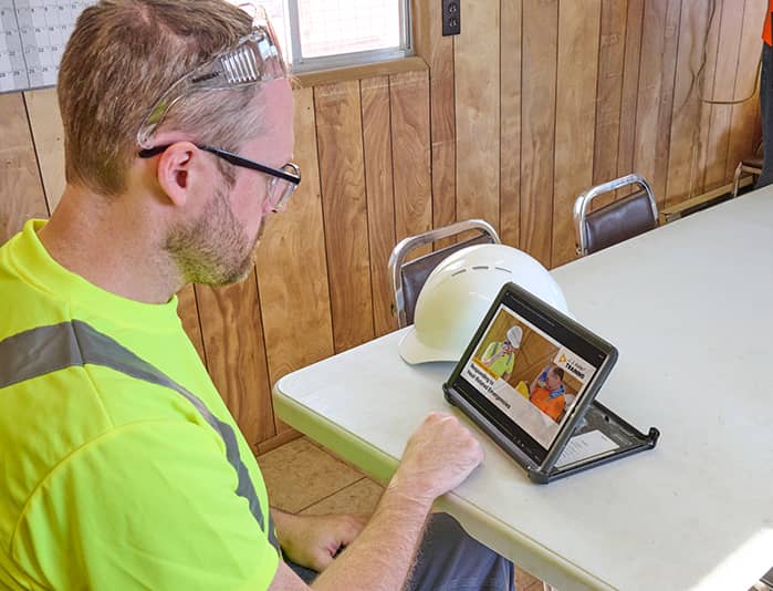 Construction worker in high-visibility gear engages in remedial corrective action training for construction on a tablet, with a hard hat on the table.