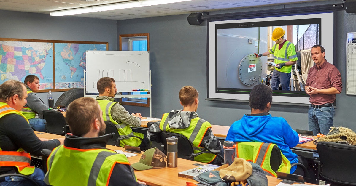 Participants wearing high-visibility safety vests are seated classroom-style while an instructor presents a training OSHA EHS video on a projection screen